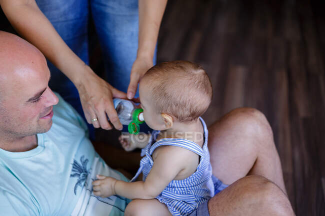 From above baby drinking water from bottle in mother hands while sitting on lap of bald father at home — Stock Photo