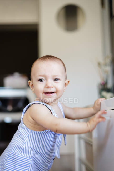 Bebê descalço excitado olhando para longe enquanto sentado em parquet perto de balcões na cozinha acolhedora em casa — Fotografia de Stock