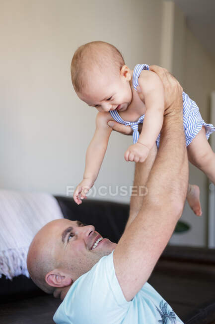 Hombre calvo encantado sonriendo y levantando al bebé alegre mientras está sentado cerca del sofá en casa - foto de stock
