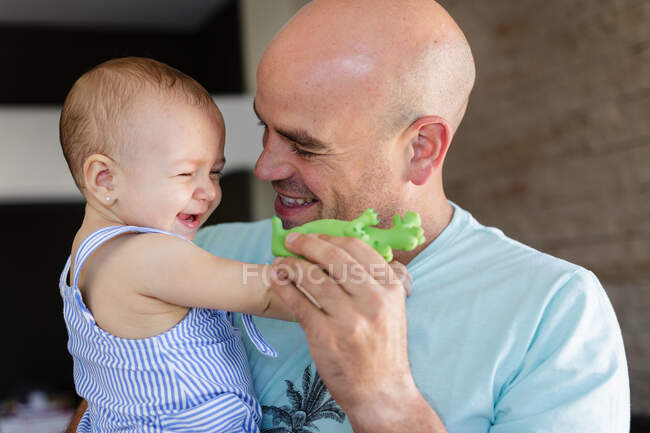 Feliz hombre calvo con juguete verde abrazándose y jugando con el bebé riendo mientras pasan tiempo en casa juntos - foto de stock