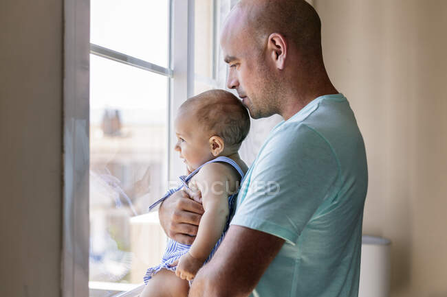 Vue latérale de l'homme chauve embrassant et embrassant bébé heureux tout en se tenant près de la fenêtre dans une chambre confortable à la maison — Photo de stock
