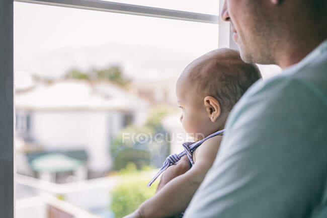 Вид збоку лисий чоловік обіймає і цілує щасливу дитину, стоячи біля вікна в затишній кімнаті вдома — стокове фото