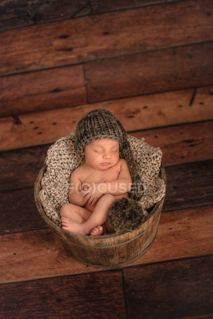 Nu bébé en bonnet tricoté dormir dans un seau sur le sol en bois à la maison — Photo de stock