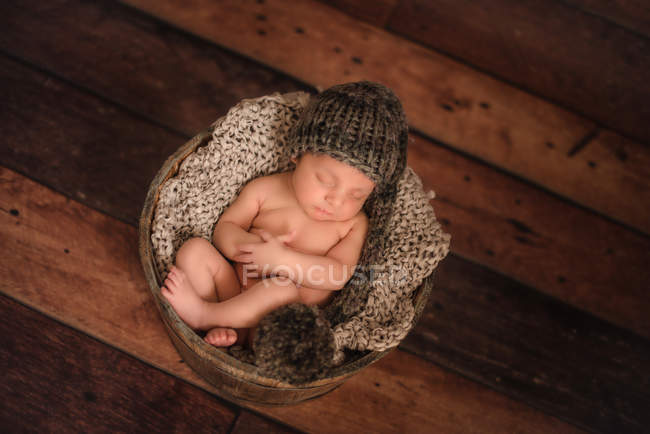 From above nude infant in knitted hat sleeping in bucket on wooden floor at home — Stock Photo