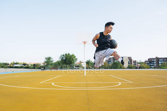 Young man jumping with black ball on yellow basketball court outdoors. — Stock Photo