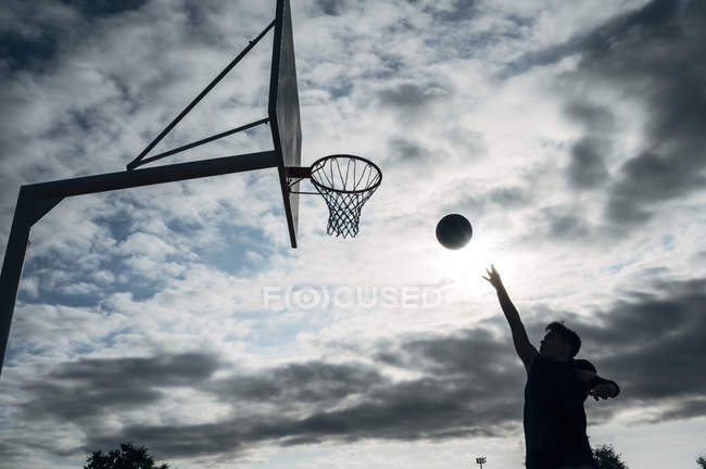 Joven saltando por anotar en cancha de baloncesto al aire libre contra el cielo nublado . - foto de stock
