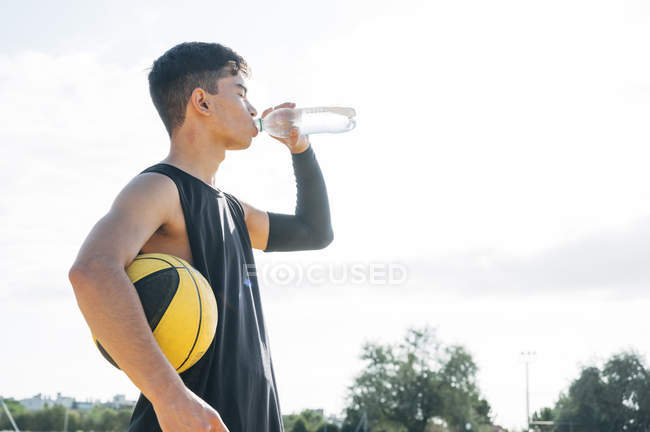 Молодой человек на баскетбольной площадке пьет воду из бутылки . — стоковое фото