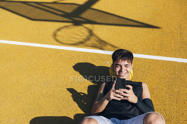 Jugador de baloncesto masculino usando smartphone mientras está acostado en la cancha amarilla después de la sesión de entrenamiento
. - foto de stock