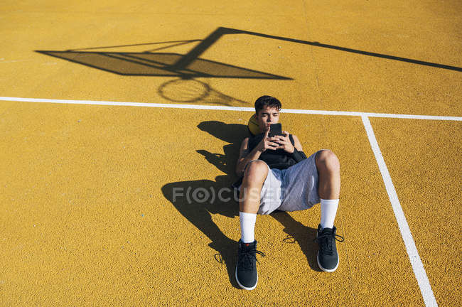 Joueur de basket masculin utilisant un smartphone tout en étant allongé sur un terrain jaune après la session d'entraînement . — Photo de stock