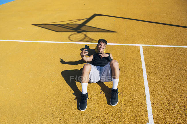 Jugador de baloncesto masculino tomando selfie con smartphone mientras está acostado en la cancha amarilla después de la sesión de entrenamiento . - foto de stock