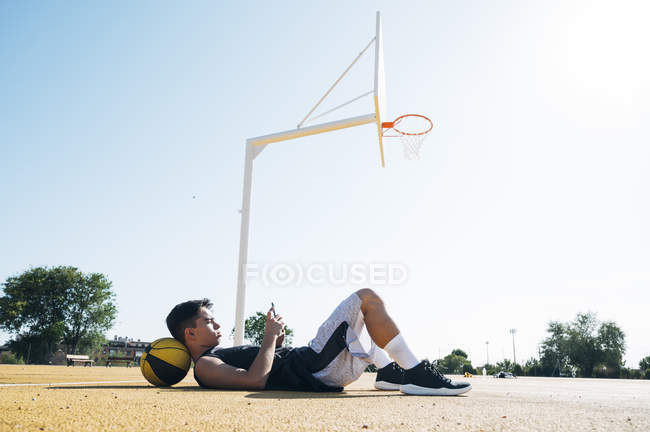 Jugador de baloncesto masculino usando smartphone mientras está acostado en la cancha amarilla después de la sesión de entrenamiento . - foto de stock