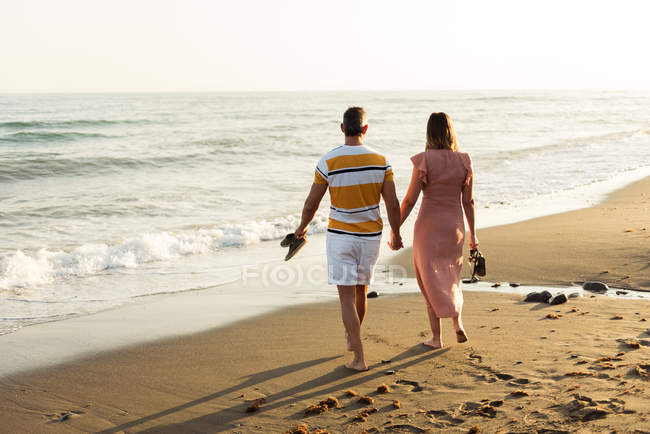 Вид збоку босоніж чоловік і жінка тримають руки і несуть взуття під час прогулянки на піщаному пляжі — стокове фото