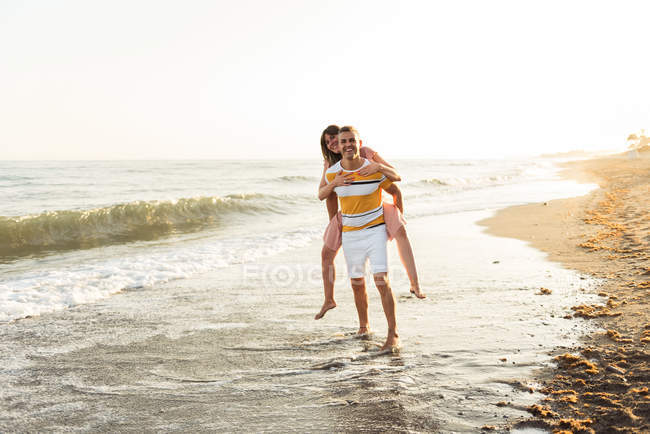 Full length cheerful male giving piggyback ride to smiling female on wet sand near waving sea — Stock Photo