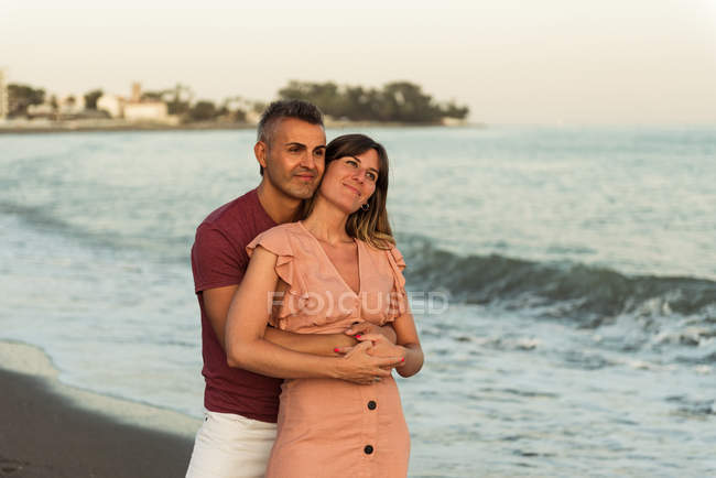 Дорослий чоловік обіймає жінку ззаду і дивиться вбік, стоячи на пляжі біля махаючого моря і відпочиваючи разом — стокове фото