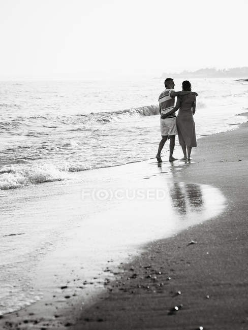 Back view of barefoot man and woman embracing while walking on sandy beach towards waving sea on resort — Stock Photo