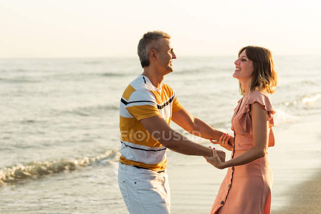 Взрослый мужчина улыбается и вращается женщина в танце, веселясь на песчаном пляже у моря — стоковое фото