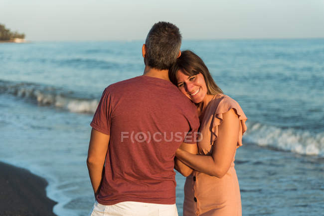Erwachsene Frau stützt sich auf Mann, während sie am Strand in der Nähe des winkenden Meeres steht — Stockfoto
