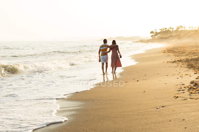 Back view of barefoot man and woman embracing and carrying shoes while walking on sandy beach — Stock Photo
