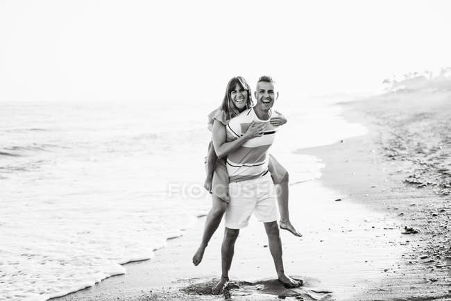 Longitud completa alegre macho adulto dando paseo a cuestas a la hembra sonriente mientras está de pie sobre arena húmeda - foto de stock