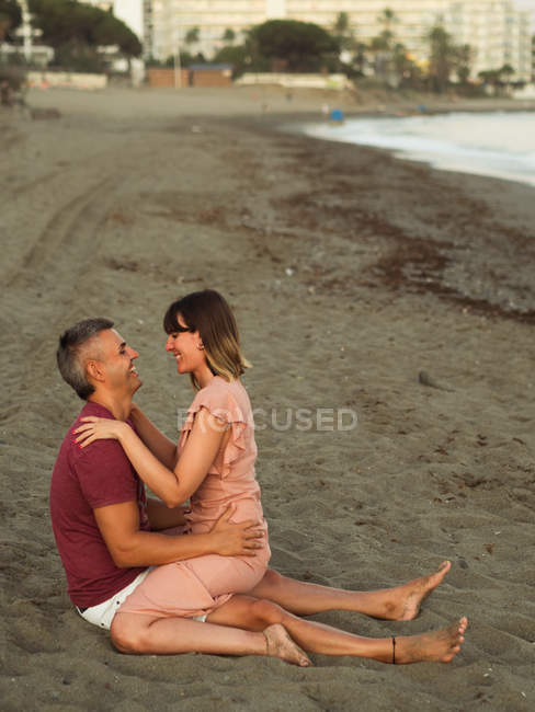 Full length adult man and woman smiling and hugging while sitting on sand near sea and relaxing during date — Stock Photo