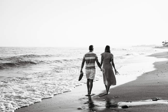 Back view of barefoot man and woman holding hands and carrying shoes while walking on sandy beach — Stock Photo