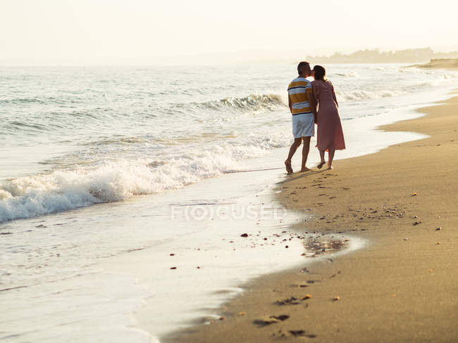 Rückansicht des barfüßigen Mannes und der barfüßigen Frau, die sich küssen, während sie am Sandstrand in Richtung des winkenden Meeres am Urlaubsort gehen — Stockfoto