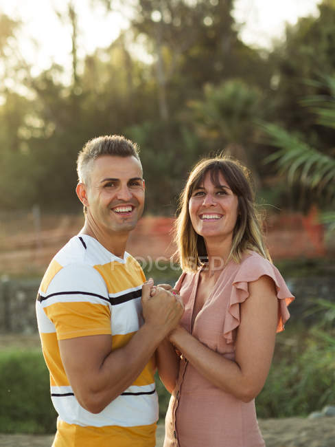 Vista lateral del hombre y la mujer adultos tomados de la mano y mirando en la cámara con sonrisa feliz - foto de stock