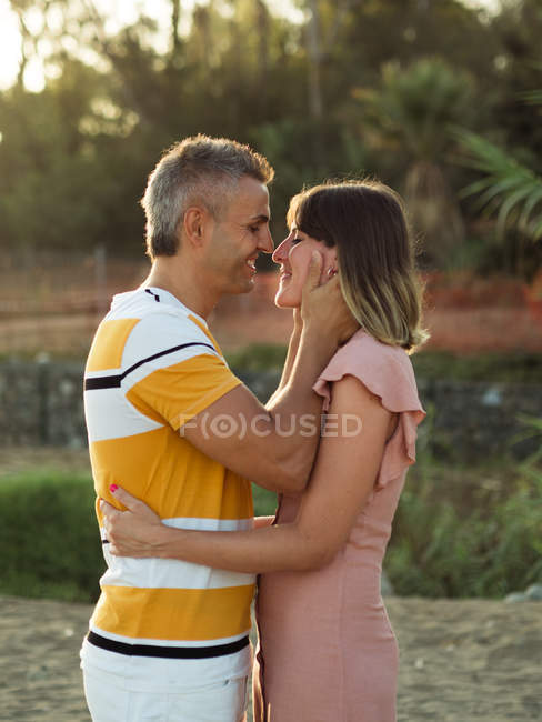 Взгляд сбоку взрослых мужчины и женщины, глядящих друг на друга со счастливой улыбкой, стоя на песчаном берегу курорта — стоковое фото