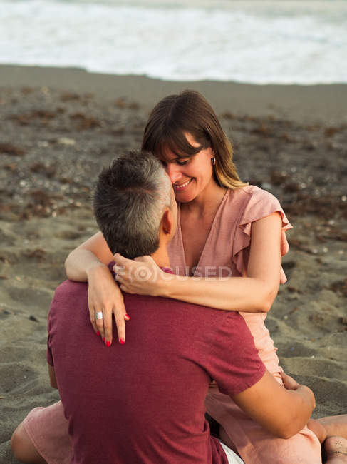 Hombre y mujer sonriendo y abrazándose mientras están sentados en la arena cerca del mar y relajarse durante la fecha - foto de stock