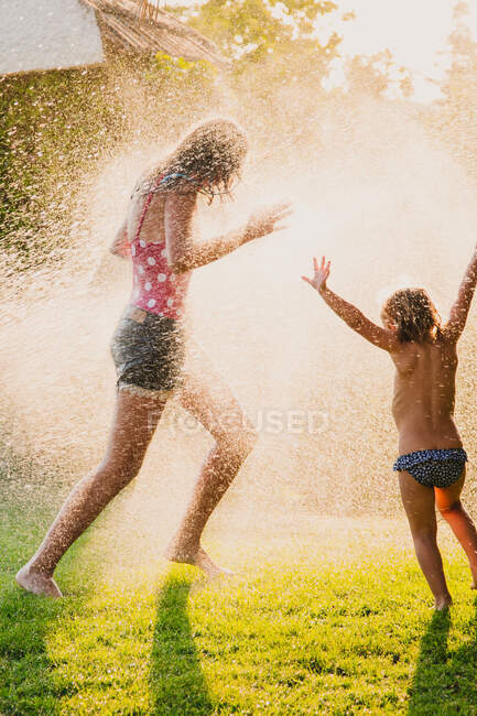 Cuerpo completo adolescente anónimo y niña corriendo y jugando en gotas de rociar agua limpia mientras se divierten en el jardín juntos - foto de stock
