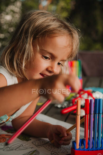 Сосредоточенная маленькая девочка, опирающаяся на стол и раскрашивающие картинки у книги маркером на размытом фоне комнаты дома — стоковое фото