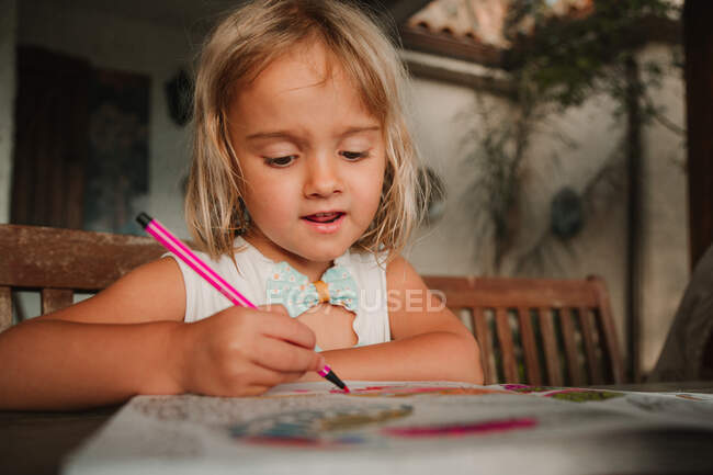 Орієнтована маленька дівчинка спирається на стіл і розмальовує картинки в книзі з маркерною ручкою на розмитому фоні кімнати вдома — стокове фото