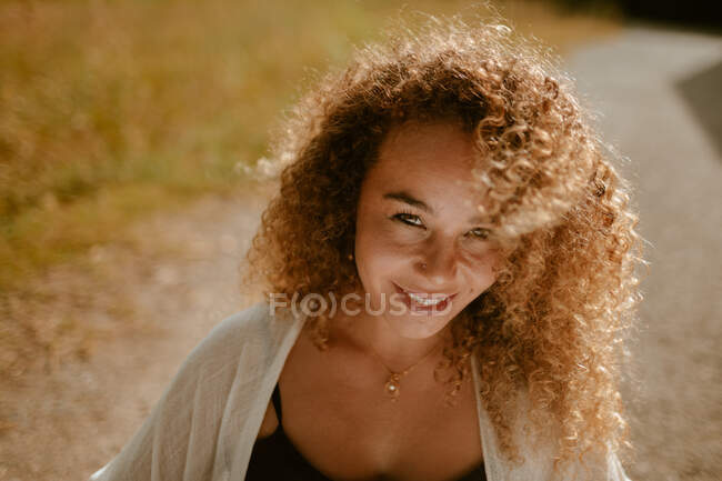 Щаслива доросла жінка з кучерявим волоссям посміхається і дивиться на камеру, стоячи на сільській дорозі в сонячний день на природі — стокове фото