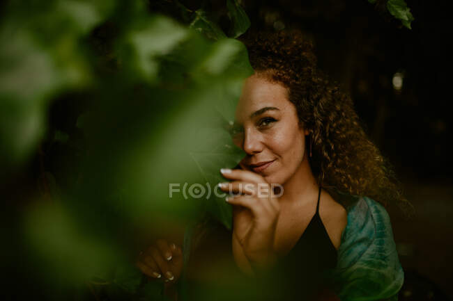 Señora adulta con el pelo rizado tocando hojas de vid verde y mirando a la cámara con sonrisa mientras pasa tiempo en el bosque oscuro - foto de stock