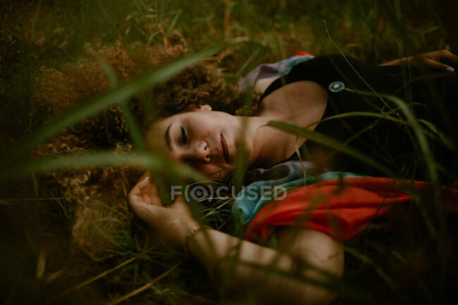 Чувственная женщина с вьющимися волосами лежит на зеленой траве и спит, проводя время в сельской местности — стоковое фото