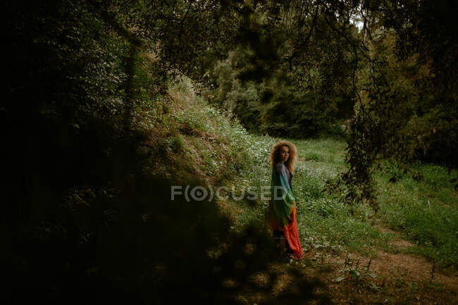 Full body side view adult female in dress looking at camera while standing near grassy hill in green forest — Stock Photo