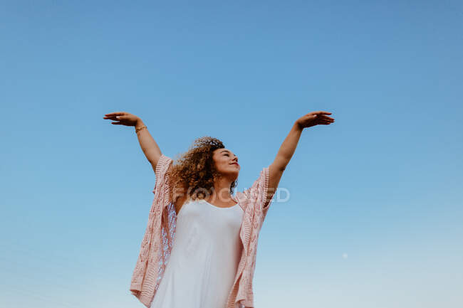 Baixo ângulo de fêmea elegante com cabelos cacheados fechando os olhos e levantando os braços enquanto está contra o céu azul sem nuvens na natureza — Fotografia de Stock