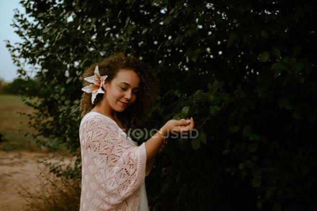 Mulher feliz com flor no cabelo encaracolado fechando os olhos e tocando folhas verdes de arbusto na natureza — Fotografia de Stock