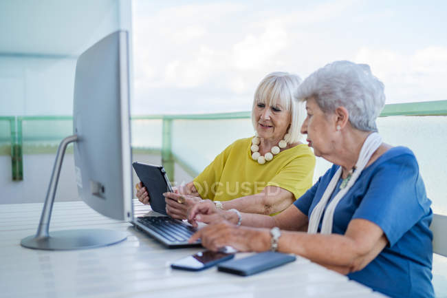 Стильні старі друзі, які переглядають соціальні медіа на планшеті, сидячи за столом з комп'ютером на балконі на курорті — стокове фото