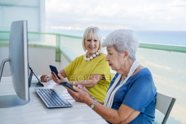 Elegantes viejos amigos navegando por las redes sociales en la tableta mientras están sentados en la mesa con el ordenador en el balcón en el resort - foto de stock