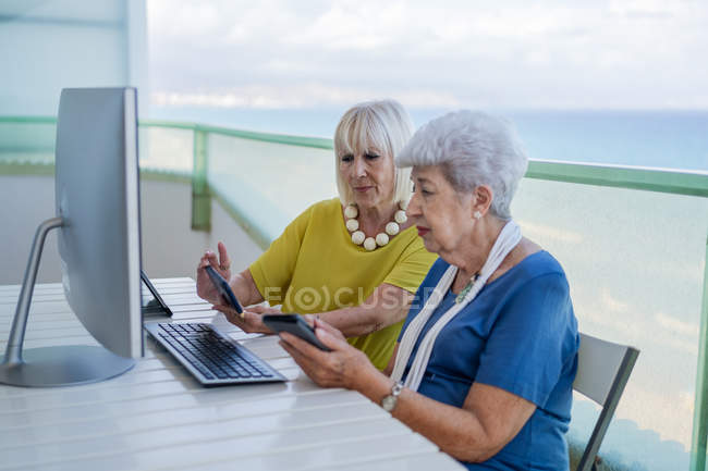 Stilvolle alte Freunde surfen auf dem Tablet in den sozialen Medien, während sie am Tisch mit dem Computer auf dem Balkon des Resorts sitzen — Stockfoto