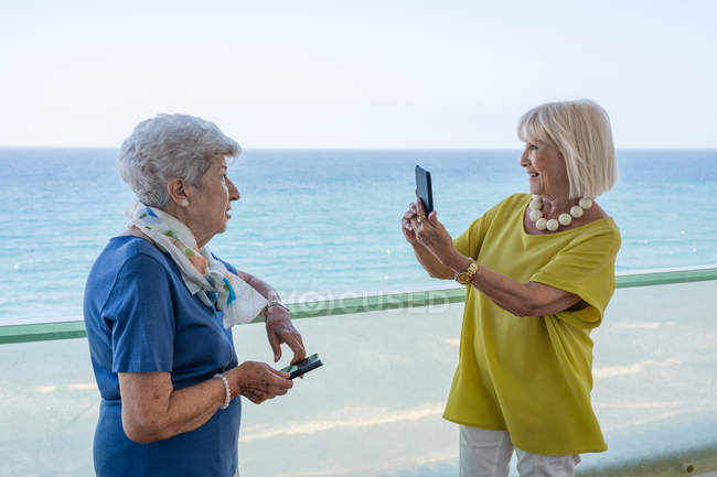 Cheerful women in stylish outfit smiling and taking picture of old friend while standing on terrace near waving sea on resort — Stock Photo