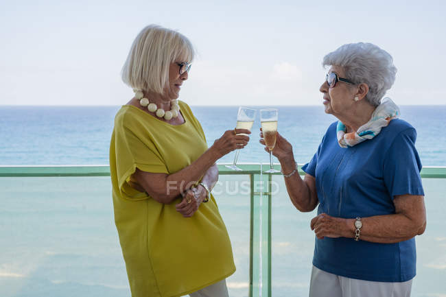 Mujeres felices en trajes elegantes diciendo brindis y disfrutando del vino mientras están de pie en el balcón del hotel cerca del mar en el resort - foto de stock
