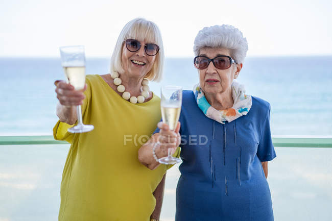 Happy women in elegant outfits saying toast and enjoying wine while standing on hotel balcony near sea on resort — Stock Photo