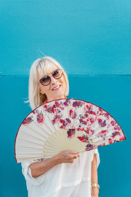 Elegante ältere Frau mit einem Fan, der lächelt und in die Kamera blickt, während er an einem heißen Tag im Resort vor einer blauen Wand steht — Stockfoto