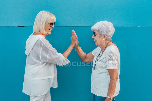 Вид сбоку смеющихся современных весёлых пожилых седых женщин в солнечных очках, дающих пять — стоковое фото