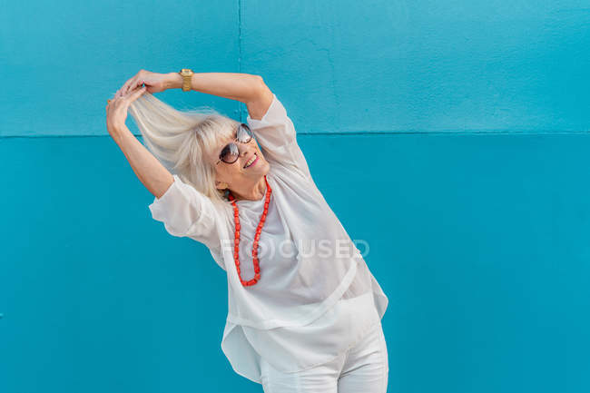 Portrait of beautiful stylish white-haired senior woman with sunglasses in white shirt on blue wall background — Stock Photo