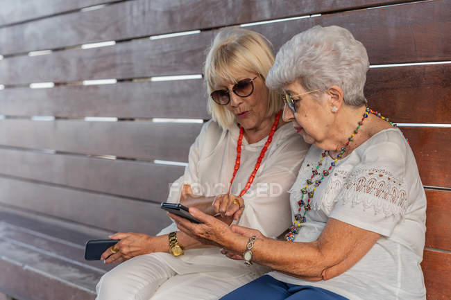 Femmes âgées aux cheveux gris gaies et à la mode et confiantes en vêtements d'été et lunettes de soleil parlant sur un grand banc en bois — Photo de stock