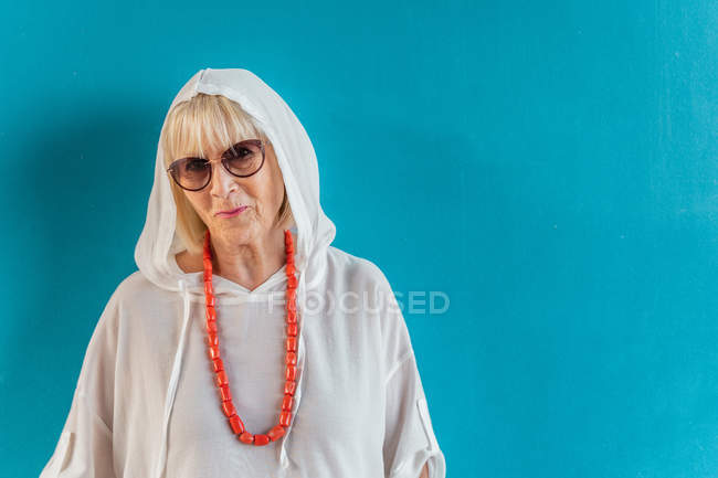 Retrato de hermosa mujer mayor de pelo blanco con gafas de sol en camisa blanca con capucha en la cabeza y cuentas de coral - foto de stock