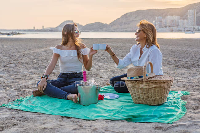 Cheerful mother and daughter smiling and looking at each other while sitting on blanket and clinking cups during family reunion picnic on beach in evening — Stock Photo